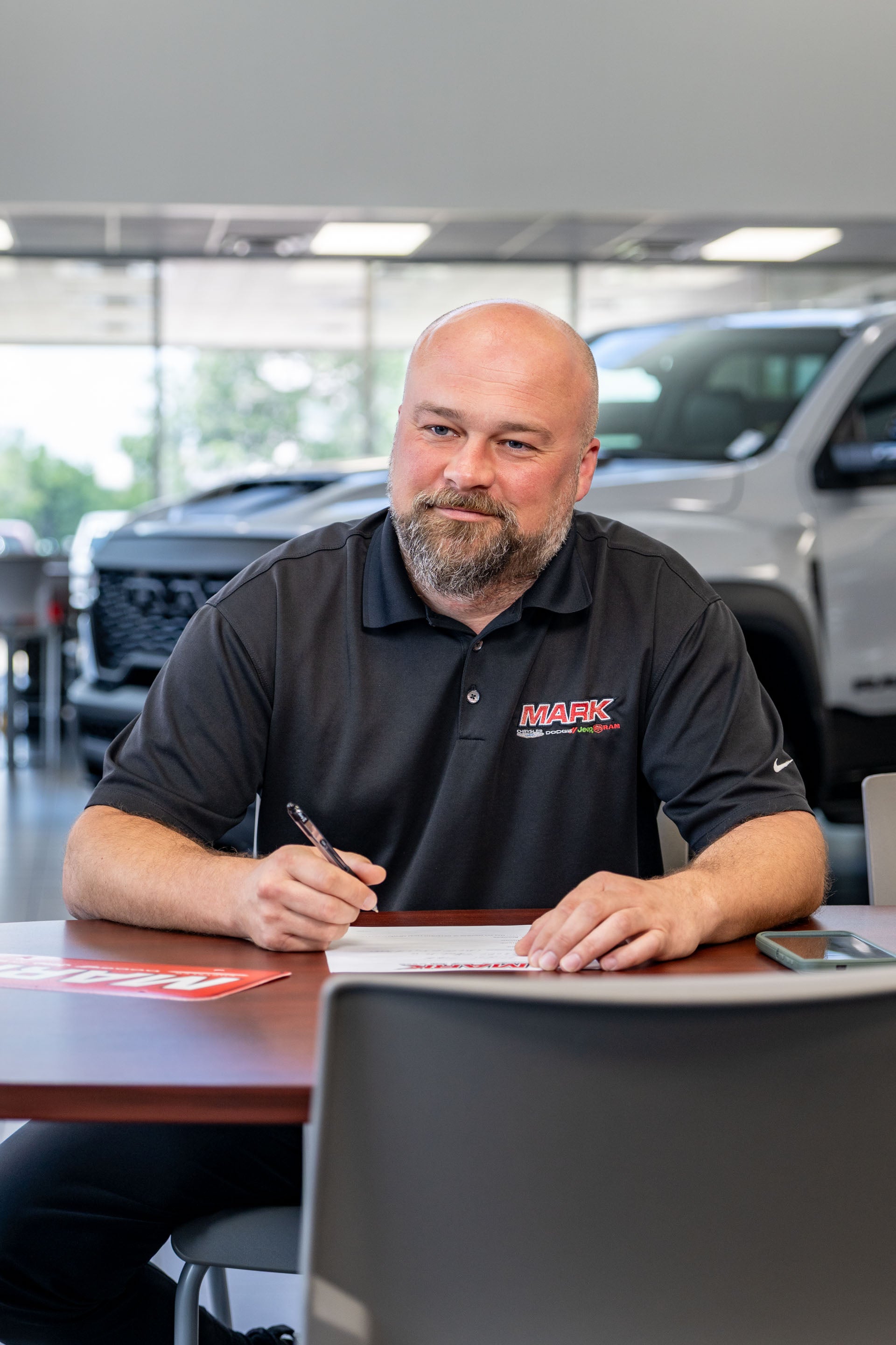 A smiling Mark Dodge staff member in a black MARK polo sits at a table writing, with a truck visible in the showroom behind him.