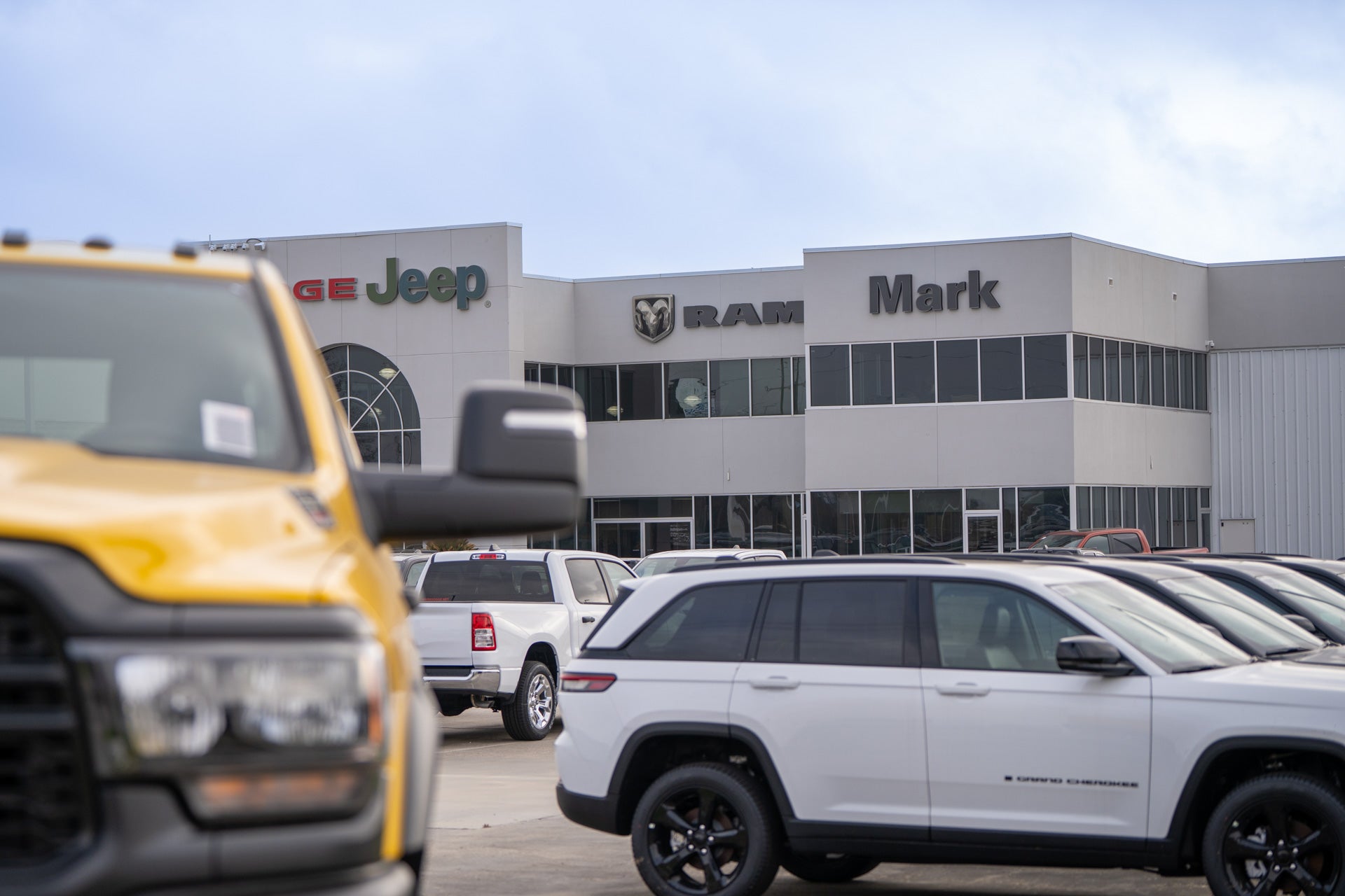 Wide exterior view of the Mark Dodge Chrysler Jeep Ram dealership building with a yellow truck in foreground and white Ram pickup and Jeep Grand Cherokee parked in the lot.