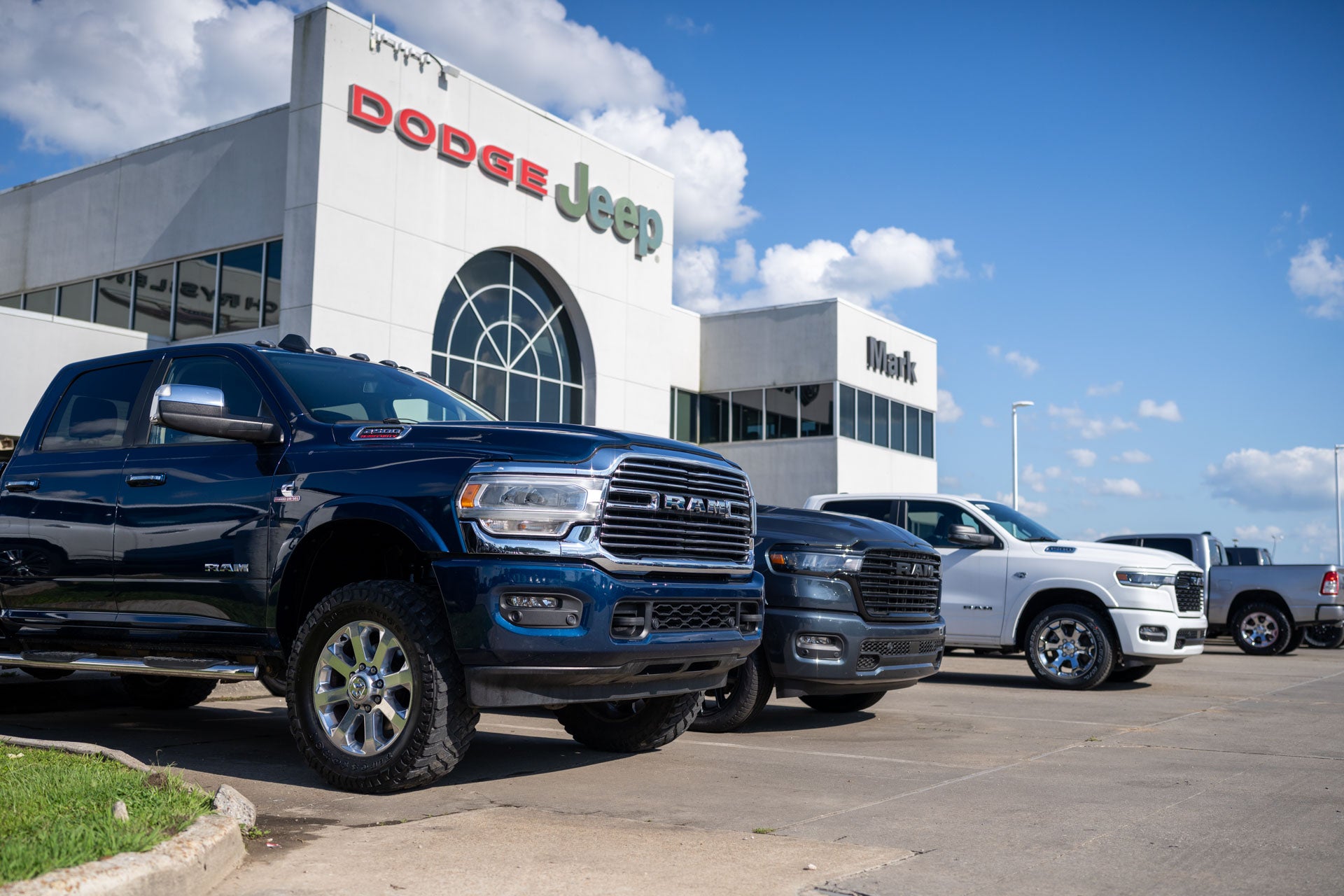 A row of Ram pickup trucks (dark blue, gray, white) parked in front of the Mark Dodge Jeep dealership building.