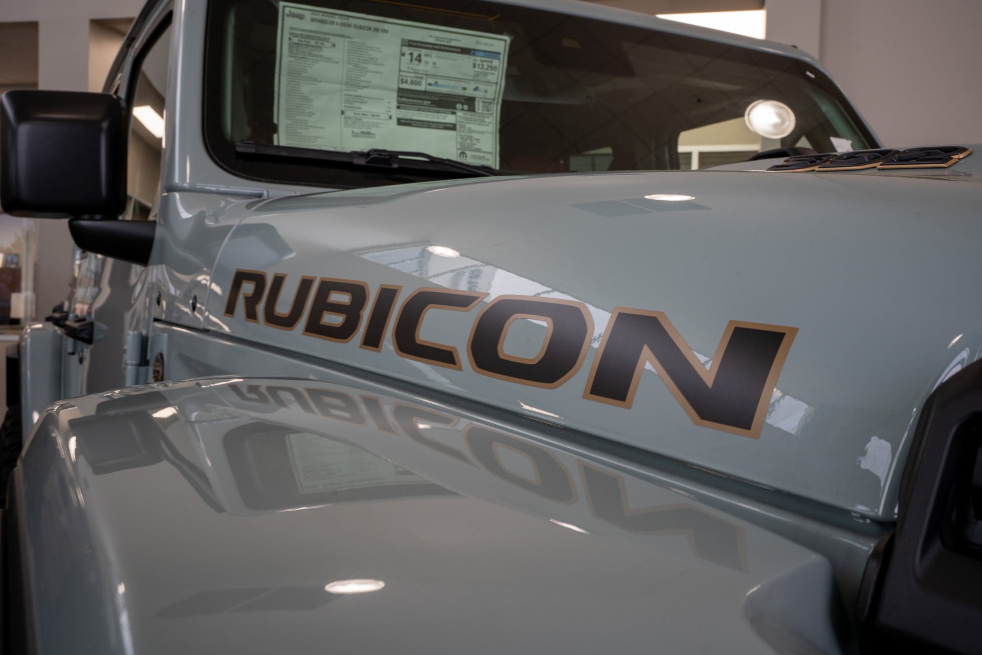Close-up of a sage green Jeep Wrangler Rubicon's hood and window sticker showing pricing, inside the Mark Dodge showroom.