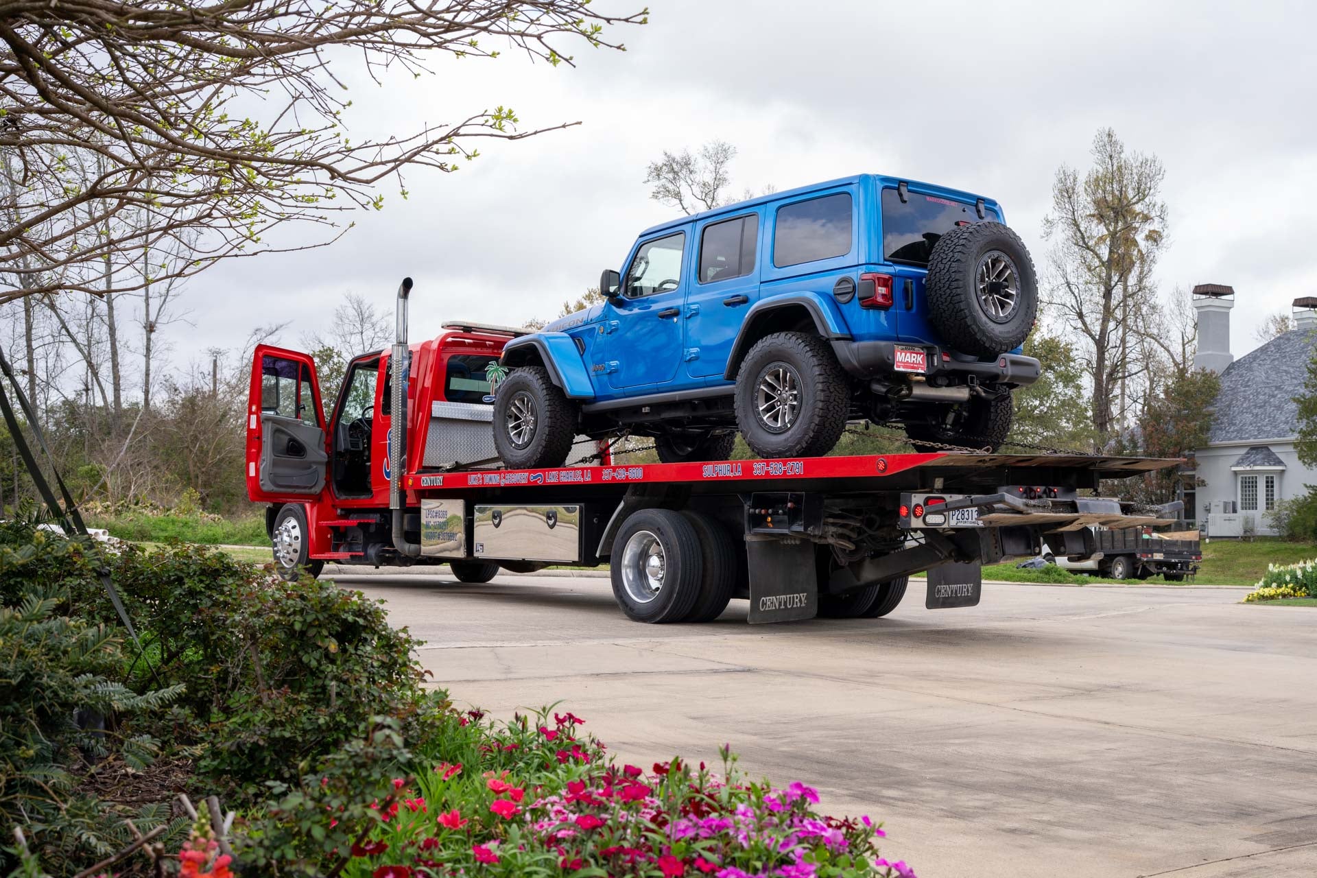 A blue Jeep Wrangler Rubicon with a Mark Dodge dealer plate being delivered on a flatbed truck, parked in a residential driveway.