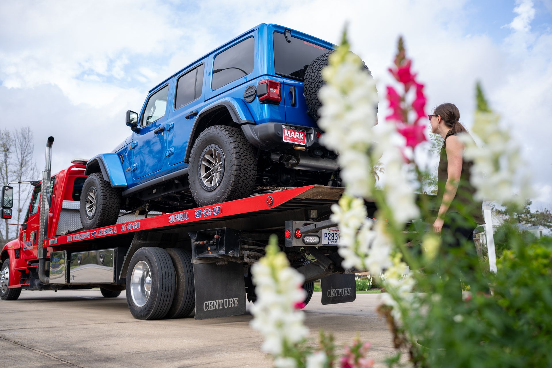 A customer in an olive top and sunglasses stands beside a blue Jeep Wrangler loaded on a flatbed truck in her driveway, framed by flowers.