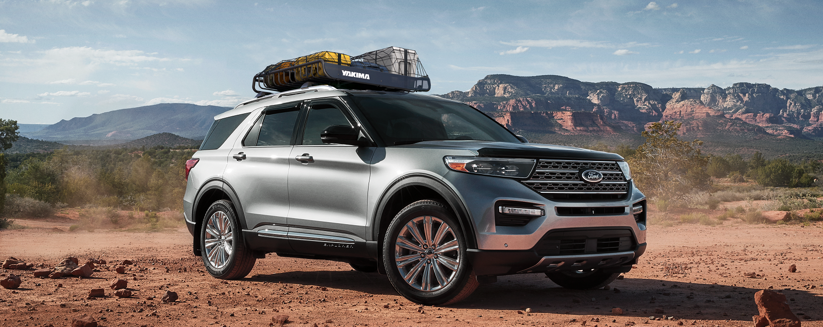 A new gray 2021 Ford Explorer with a roof rack filled with luggage in the dessert with brush and mountains pictured in the distance.