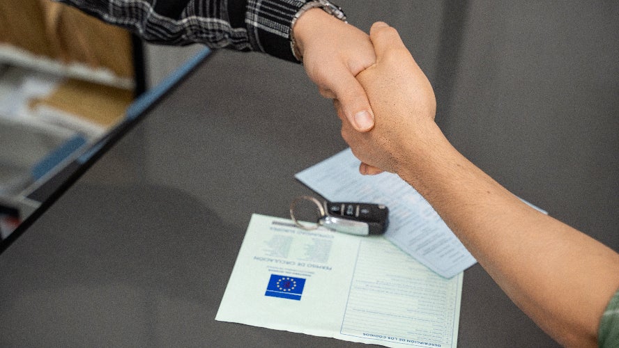 Two people shaking hands over a vehicle contract and key fob.