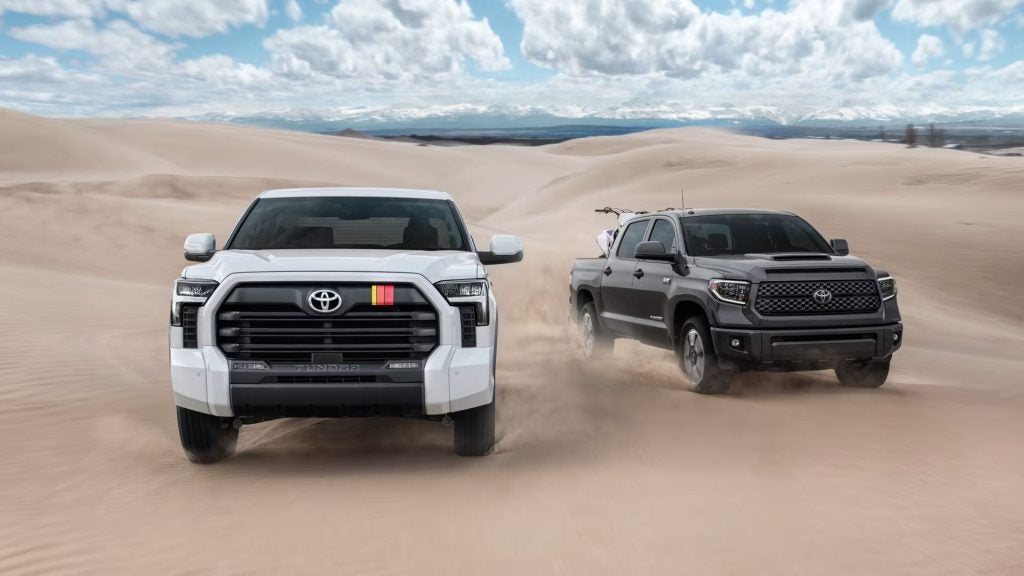 A white and a gray Toyota Tundra pickup truck kicking up sand in the desert, with distant mountains.