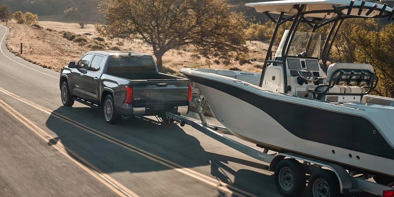A dark grey pickup truck tows a boat on a trailer up a winding road through a dry, hilly landscape.