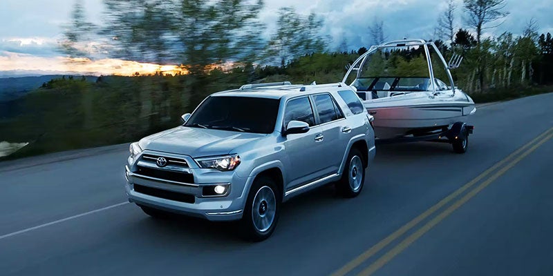 Silver Toyota 4Runner SUV towing a white motorboat on a trailer down a road at dusk.