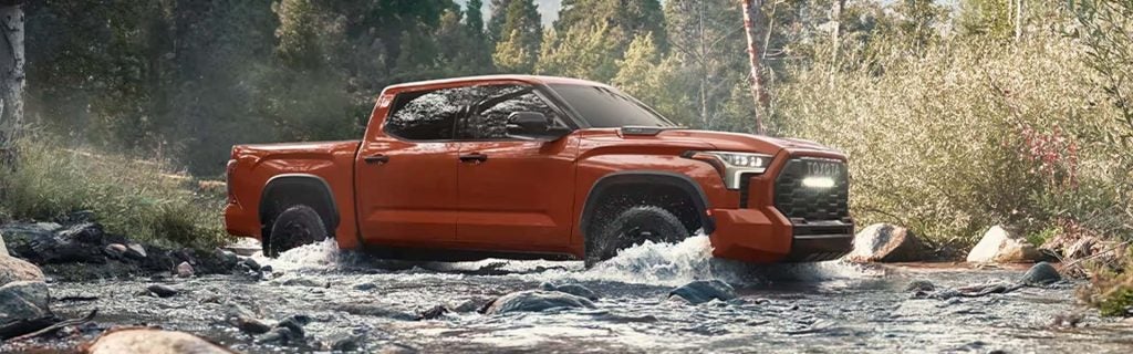 A red pickup truck drives through a rocky stream with trees and brush in the background.