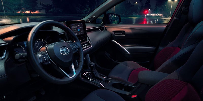 Interior shot of a car at night, showing the steering wheel, dashboard with a screen, and front seats with red accents.