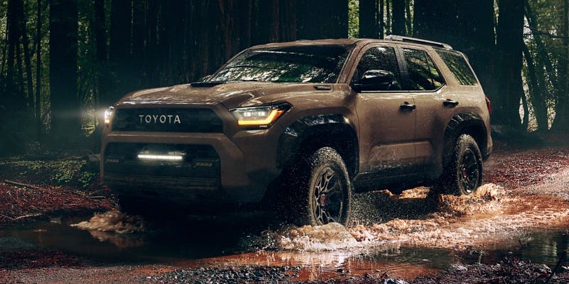 A brown Toyota SUV with its headlights on, splashing through a muddy puddle in a dark forest.