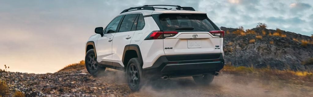Rear view of a white Toyota RAV4 with roof racks on a dirt incline, dusty and moving away.