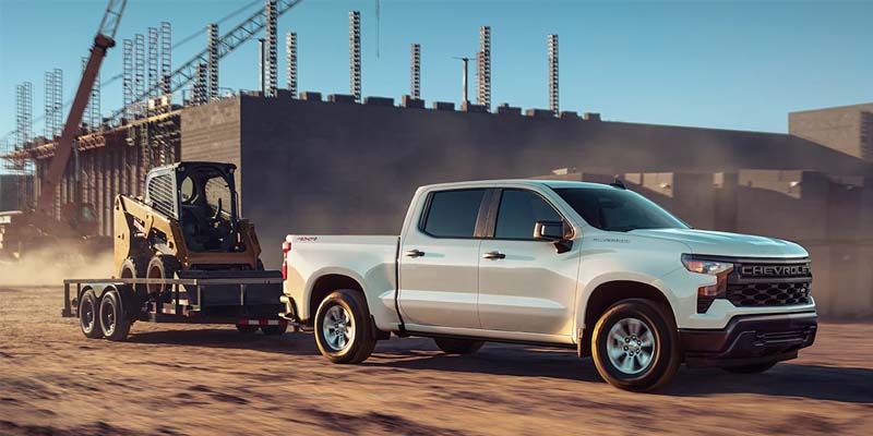 White Chevrolet Silverado pickup truck towing a yellow skid steer loader on a trailer at a dusty construction site