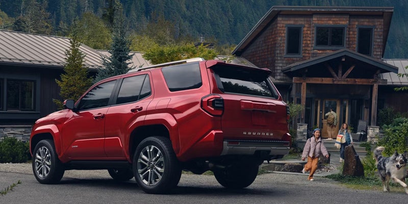 Red SUV parked on a driveway with two children, an adult, and a dog walking from a rustic house towards it.