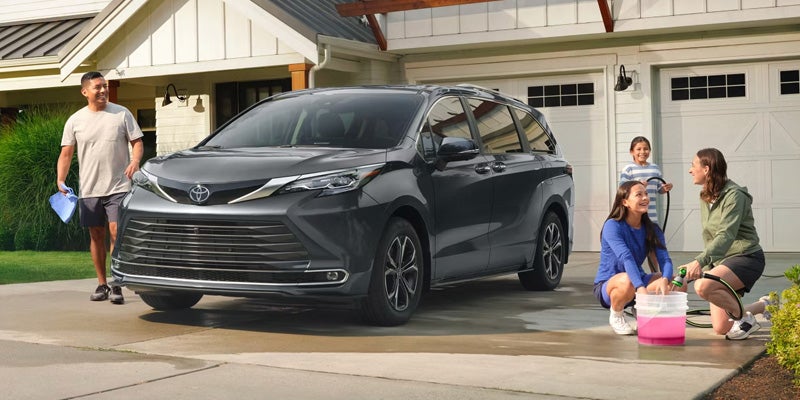 Family washing a dark gray minivan in the driveway of a light-colored house with a two-car garage.