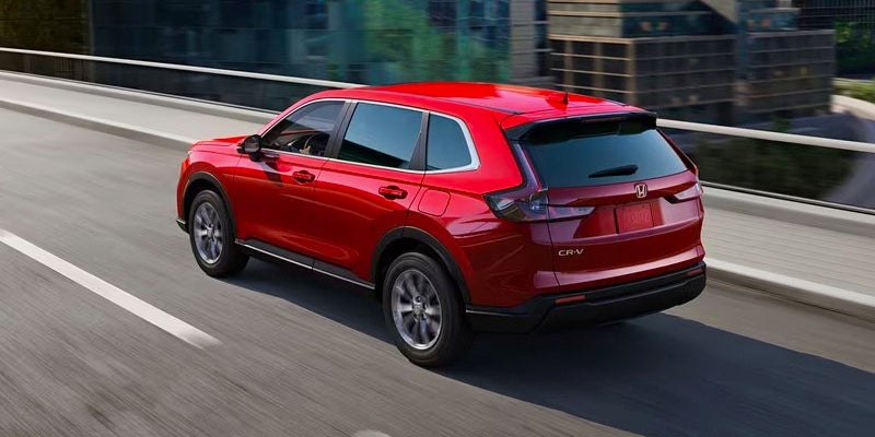 Rear view of a red Honda CR-V crossover SUV driving on a multi-lane highway, with city buildings in the background.