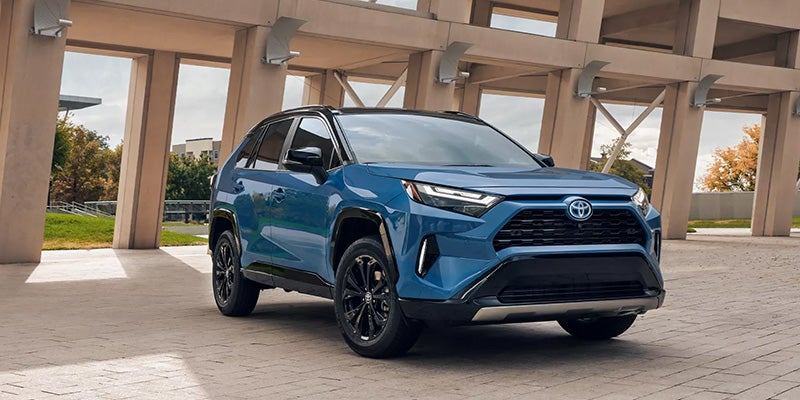 A blue Toyota RAV4 Hybrid XSE parked outdoors under a modern concrete structure, trees in background.