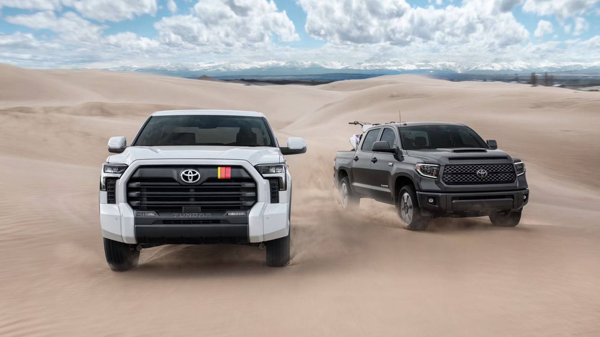 Front view of a white Toyota Tundra with a colored stripe on the grille and a gray Tundra driving on sand dunes.