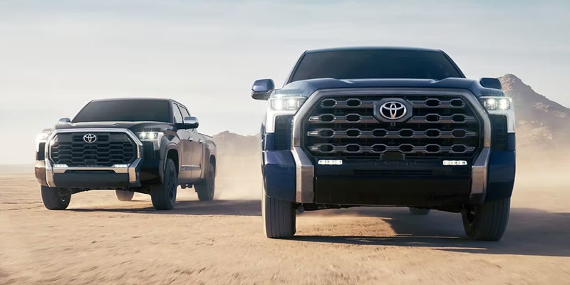 Two Toyota Tundra trucks, one blue and one black, driving side-by-side on a desert road with mountains.