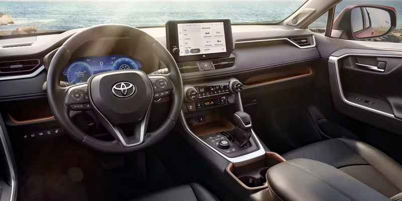 Interior view of a Toyota RAV4, showing a steering wheel, dashboard, infotainment screen, and a dark leather seat.