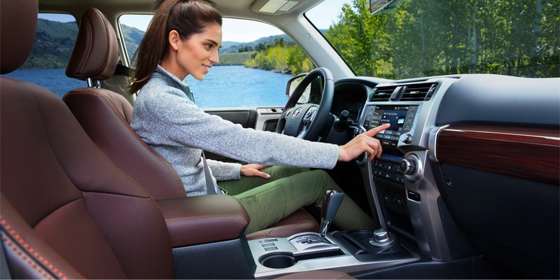 A woman in a gray sweater and green pants adjusting the touchscreen in a car with brown leather seats and a lake view.