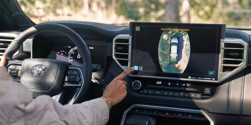 Person touching a Toyota's dashboard screen displaying a 360-degree top-down view of a car, surrounded by dirt.