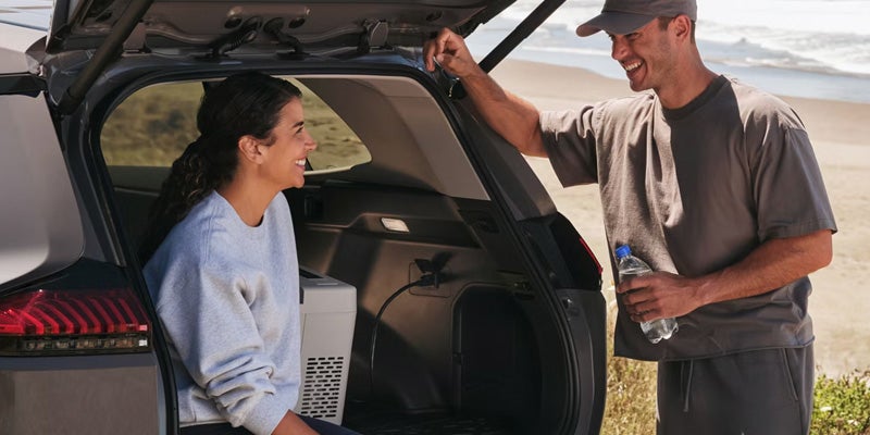 A smiling couple by the trunk of a car at the beach, with a portable cooler plugged in.