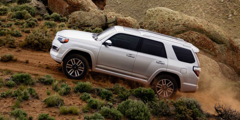 Silver Toyota 4Runner SUV climbing a dirt hill, kicking up dust, with sparse green brush and large rocks.