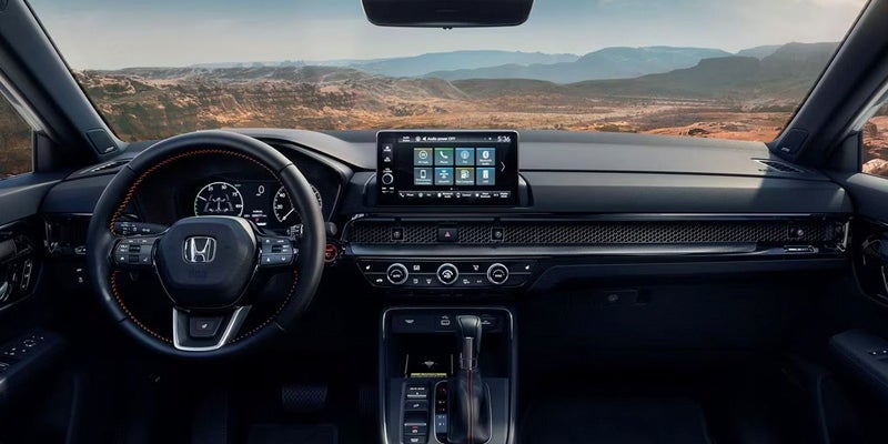 The driver's perspective inside a car, showing the dashboard, touchscreen, steering wheel, and a mountainous landscape through the windshield.