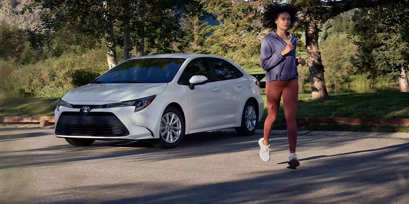 A young woman in workout clothes jogs past a white Toyota Corolla parked on a paved road, bordered by trees and a pond in the background.