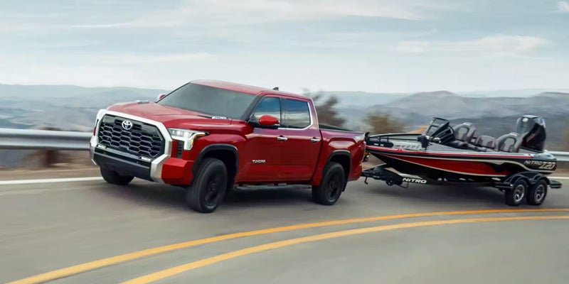 A red Toyota Tundra pickup truck towing a black and red Nitro fishing boat on a trailer, driving on a winding paved road on a cloudy day.