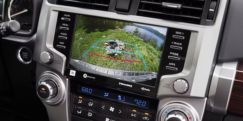 A car's dashboard screen displays a backup camera view of a campsite with a campfire pit by a lake.