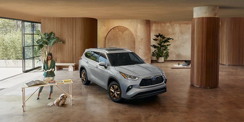 An upscale living space with a silver SUV in the center, a woman reading a book, and a dog next to a small table.