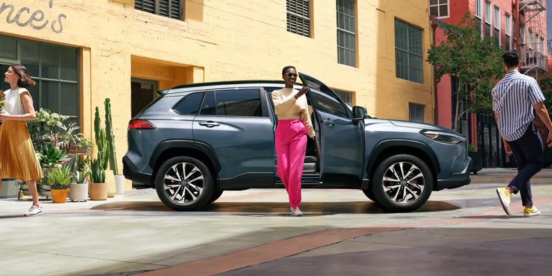 A teal SUV parked on a city street, a woman in pink pants getting out of the driver's seat.