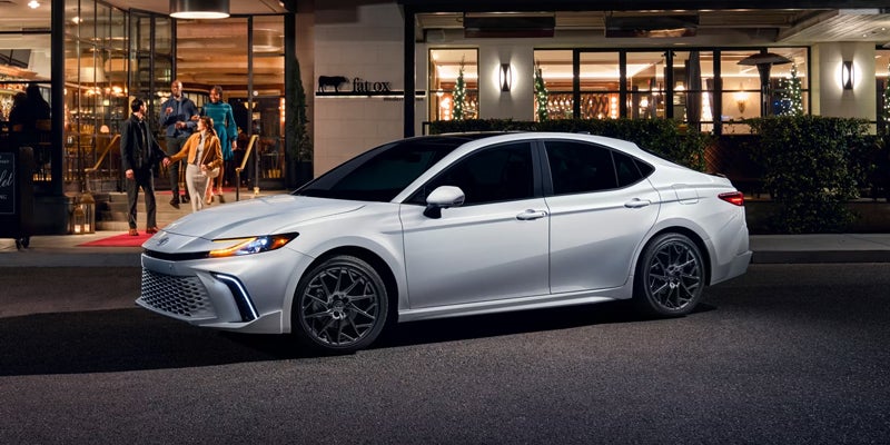 A white sedan parked at night with headlights on, in front of a restaurant with people walking out the door.