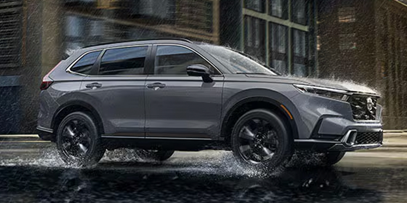 Dark gray SUV driving through a puddle on a rainy street with a building in the background.