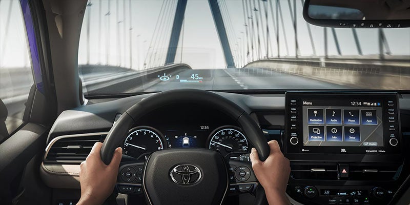 The hands on the steering wheel, showing the dashboard, infotainment screen, and a head-up display projecting speed