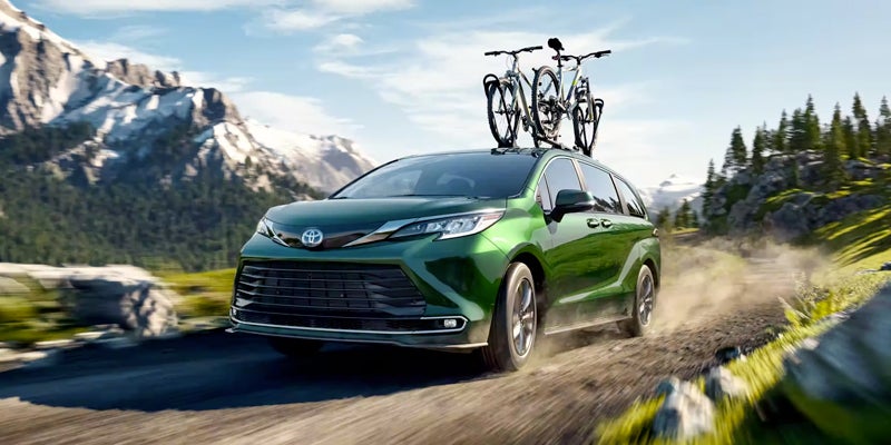 A green minivan with two bikes on its roof rack drives on a dirt road through mountains and forests.