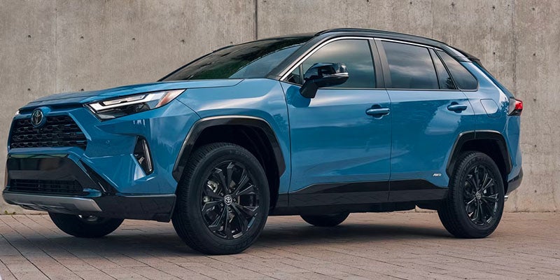 A blue Toyota RAV4 SUV with a black roof and black wheels is parked on a paved surface next to a concrete wall.