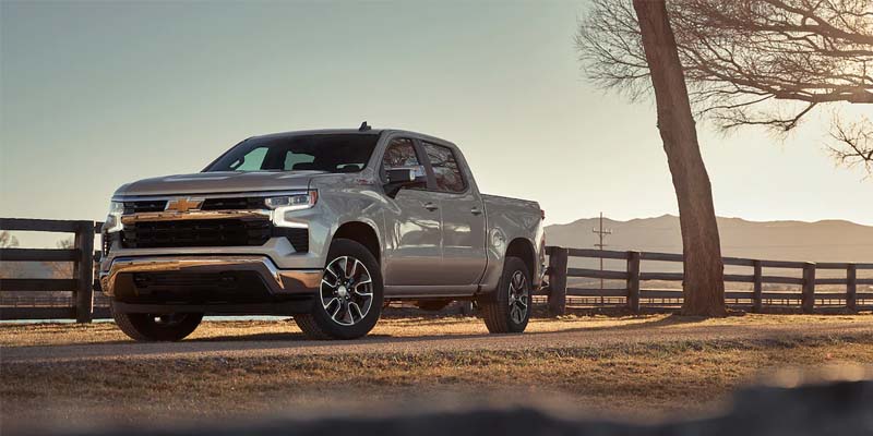 Silver pickup truck on a dirt road, with a wooden fence and tree next to it, mountains in the distance.