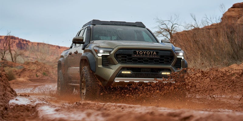 A dark green Toyota pickup truck with bright headlights drives through a muddy, unpaved road, splashing mud.