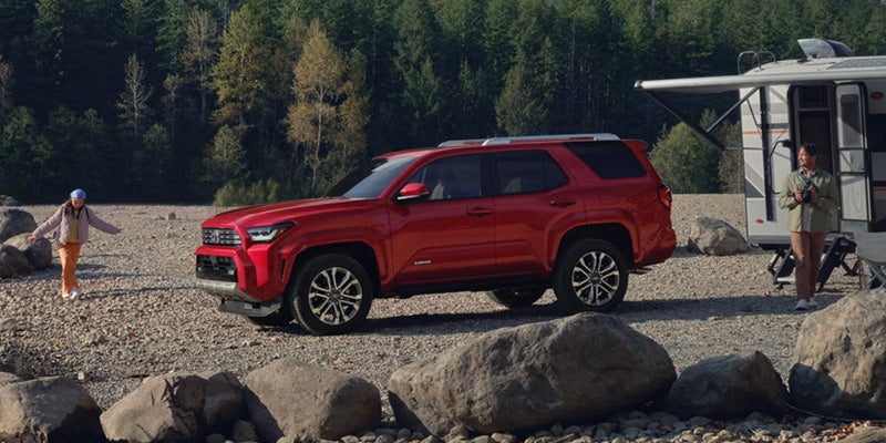 Red SUV parked on a rocky riverbank with a family, camper, and forest in the background.