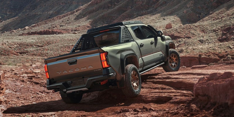 A grey-green Toyota Tacoma pickup truck with a black roll bar drives off-road in a rocky, red canyon during the day.