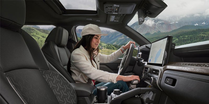 A woman in a cream jacket and hat drives a car with a mountain landscape visible through the windows.