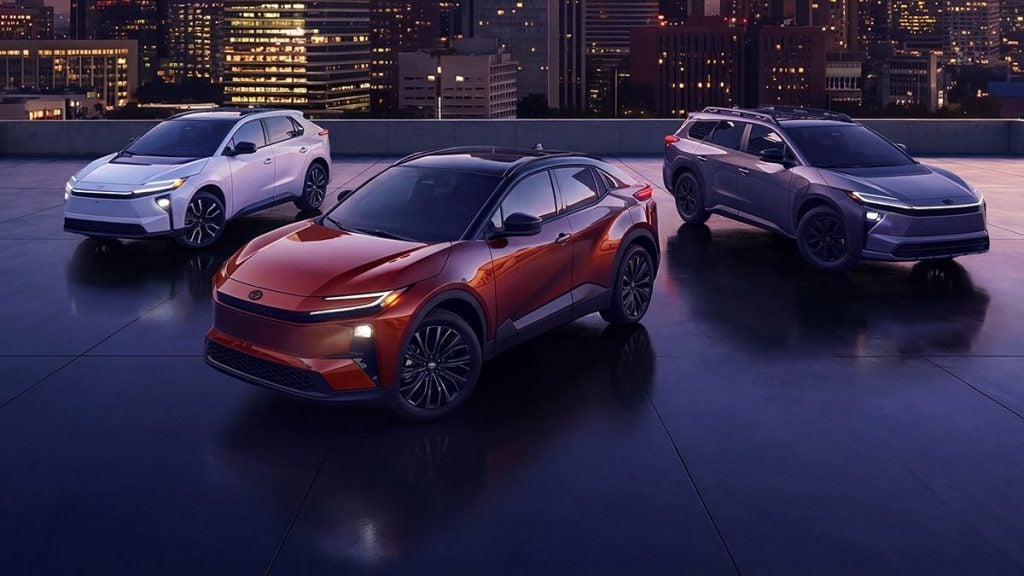 Three sleek, modern SUVs—white, burnt orange, and dark grey—parked on a rooftop at night with city lights.