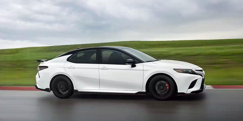 Side view of a white sports sedan with a black roof, spoiler, and trim, driving on a race track with green hills.