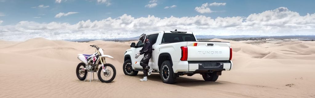 White motorcycle, rider in black gear, and white Toyota Tundra truck parked on sand