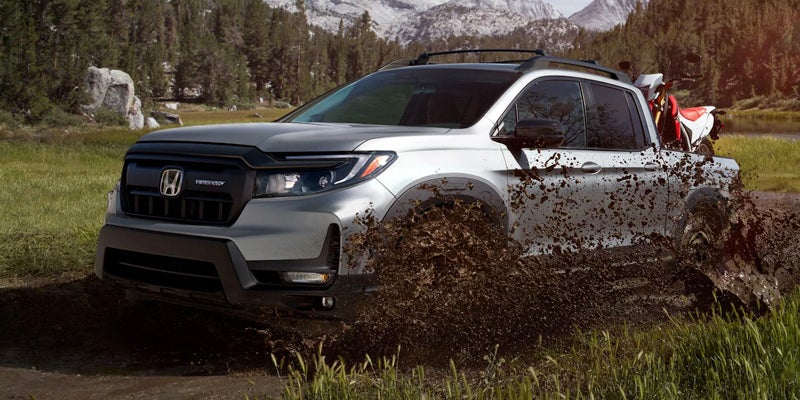 Silver pickup truck splashing through mud with a dirt bike in its bed, set against a forest and mountains