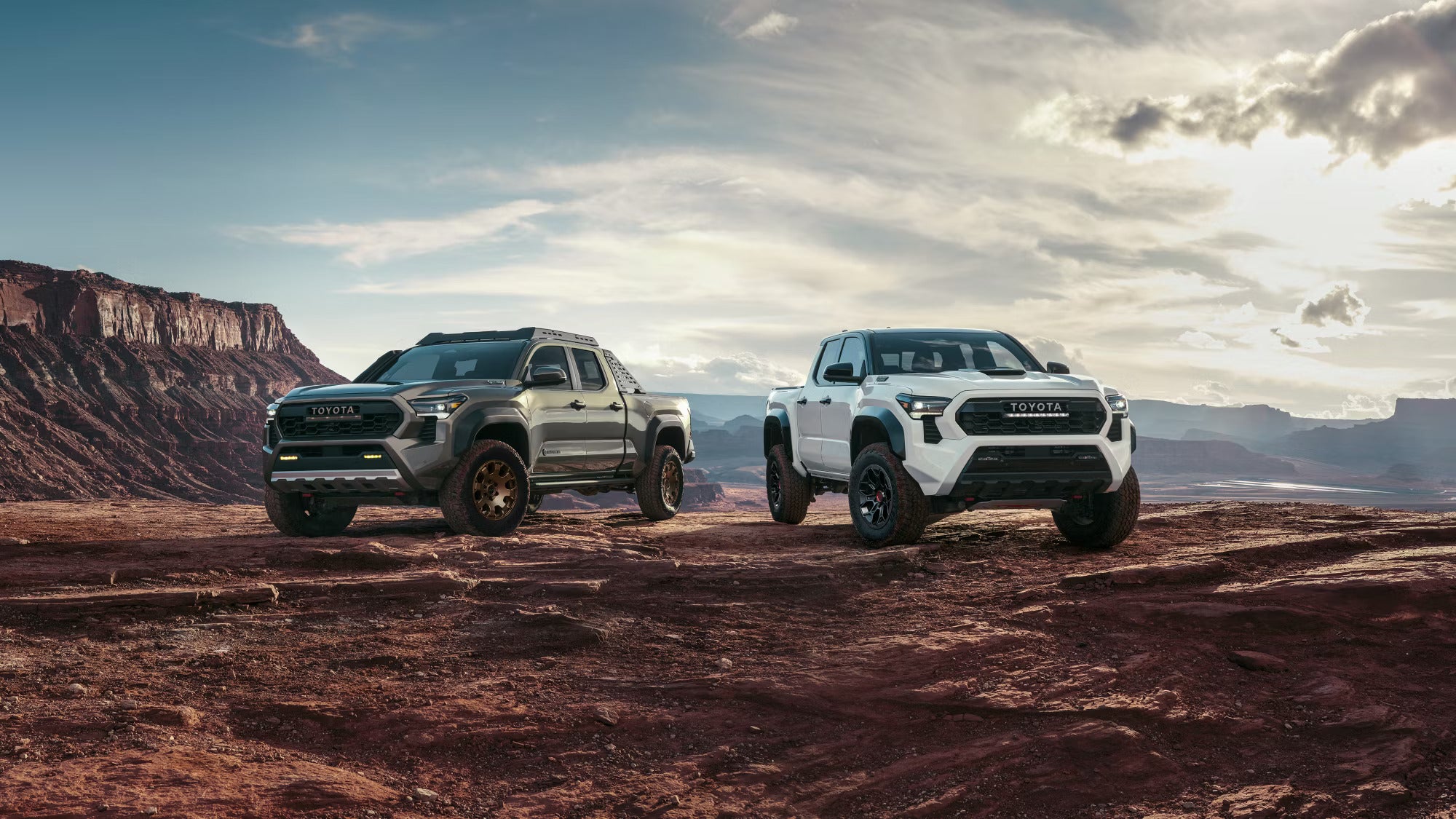 Two Toyota Tacoma pickup trucks, one gray and one white, parked on a rocky desert landscape under a cloudy sky.