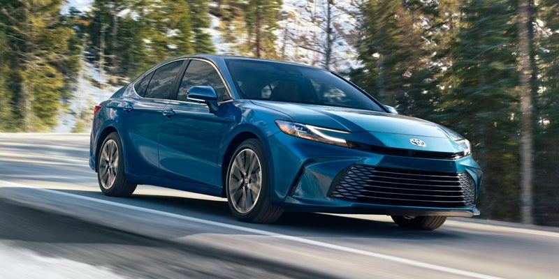 A bright teal sedan drives quickly on a winding snowy road lined with dark pine trees.