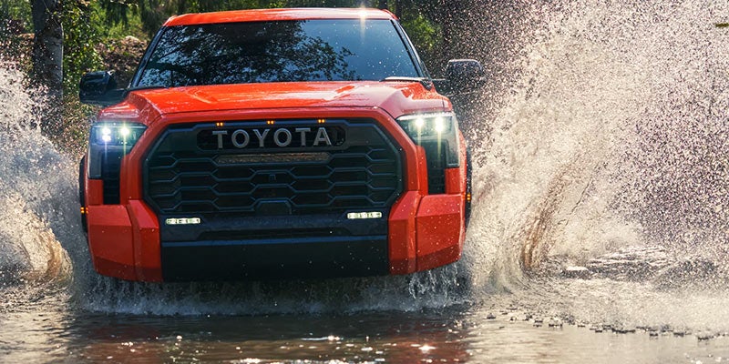 Front view of an orange Toyota truck splashing through a muddy water puddle with bright headlights on.